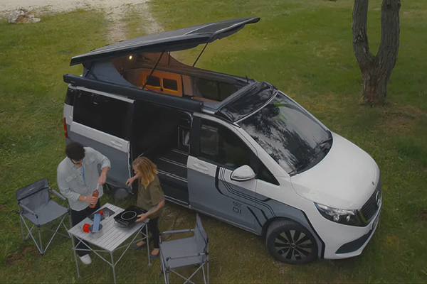 Mercedes EQV Elektro Camper mit modernem Linienmuster in Grau weiß – mit Pärchen und Tisch und Stühlen auf Gras beim Campen