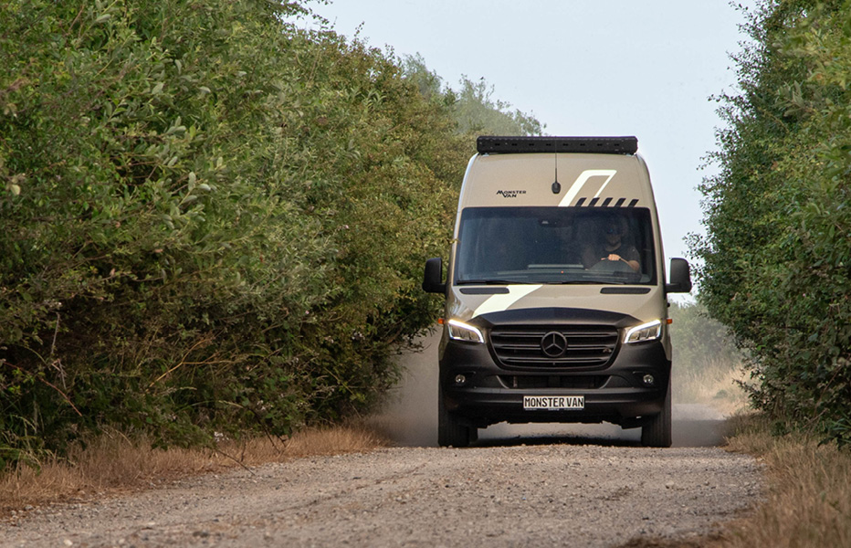 Vorderansicht von Mercedes Sprinter Camper mit schwarz/weißen Linien und sandfarbener Folierung auf Sandweg mit Büschen